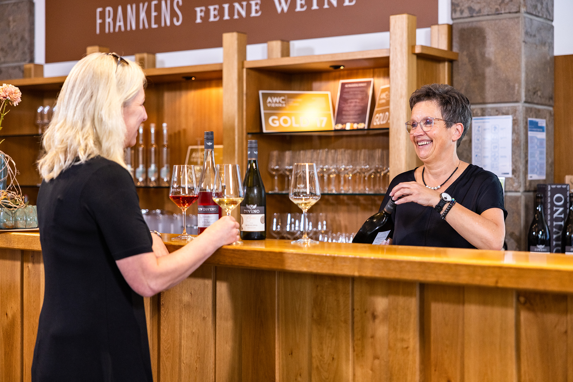 Zwei Frauen im Gespräch an der Weinbar beim Einschenken eines Glases Wein.