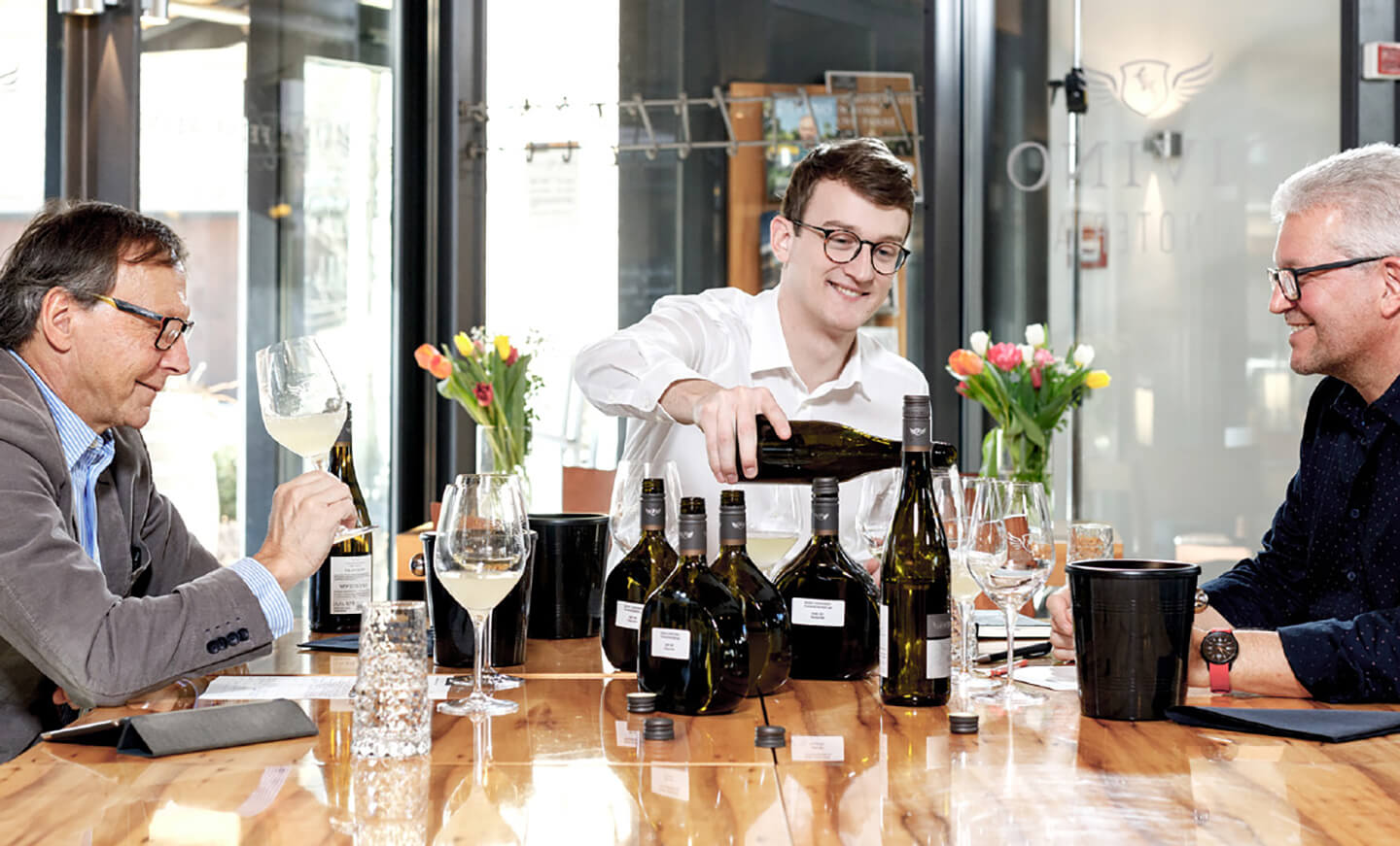 Drei Personen bei einer professionellen Weinprobe an einem Tisch mit mehreren Flaschen.